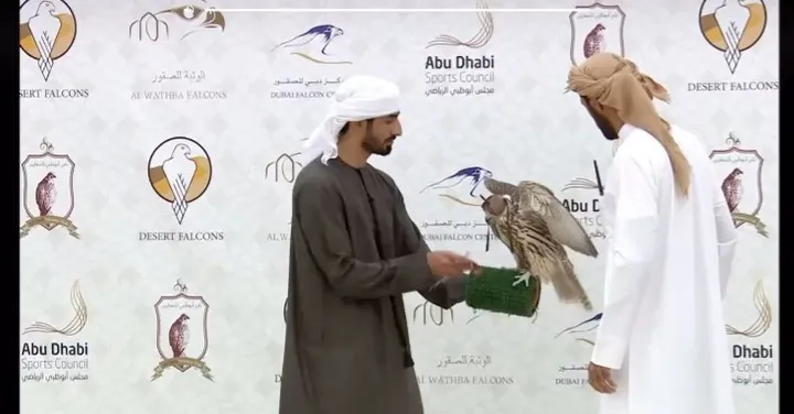 Two men in white exchanging a falcon on the fist in front of an Abu Dhabi sponsor wall